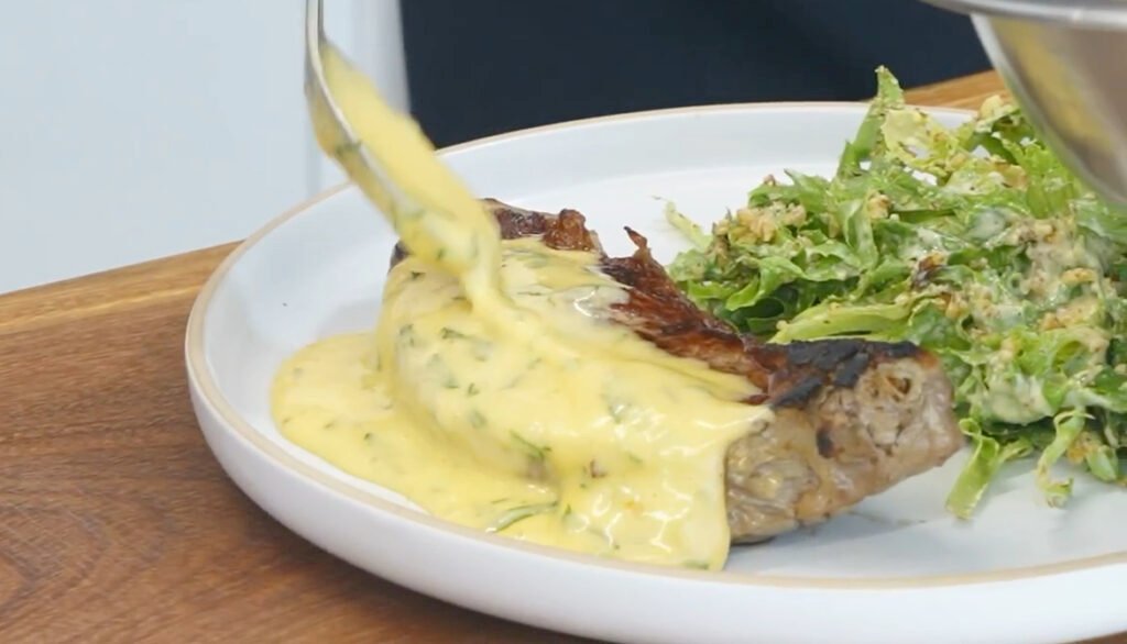 Grilled steak topped with creamy yellow cheese sauce, served with a side of green salad on a white plate.